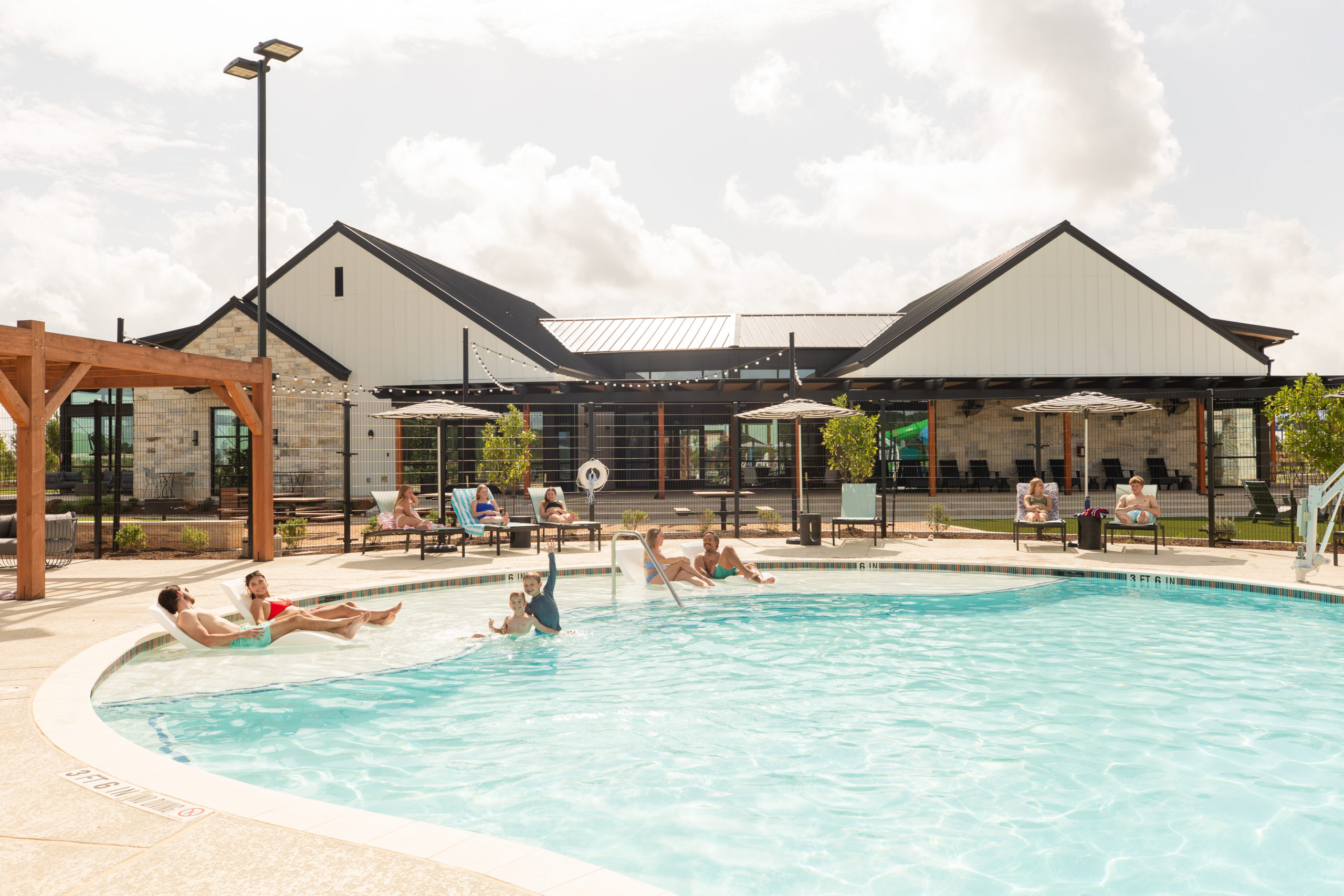 People relax and swim in an outdoor pool on a sunny day at The Haven Amenity Village, with a modern building and shaded seating areas in the background.