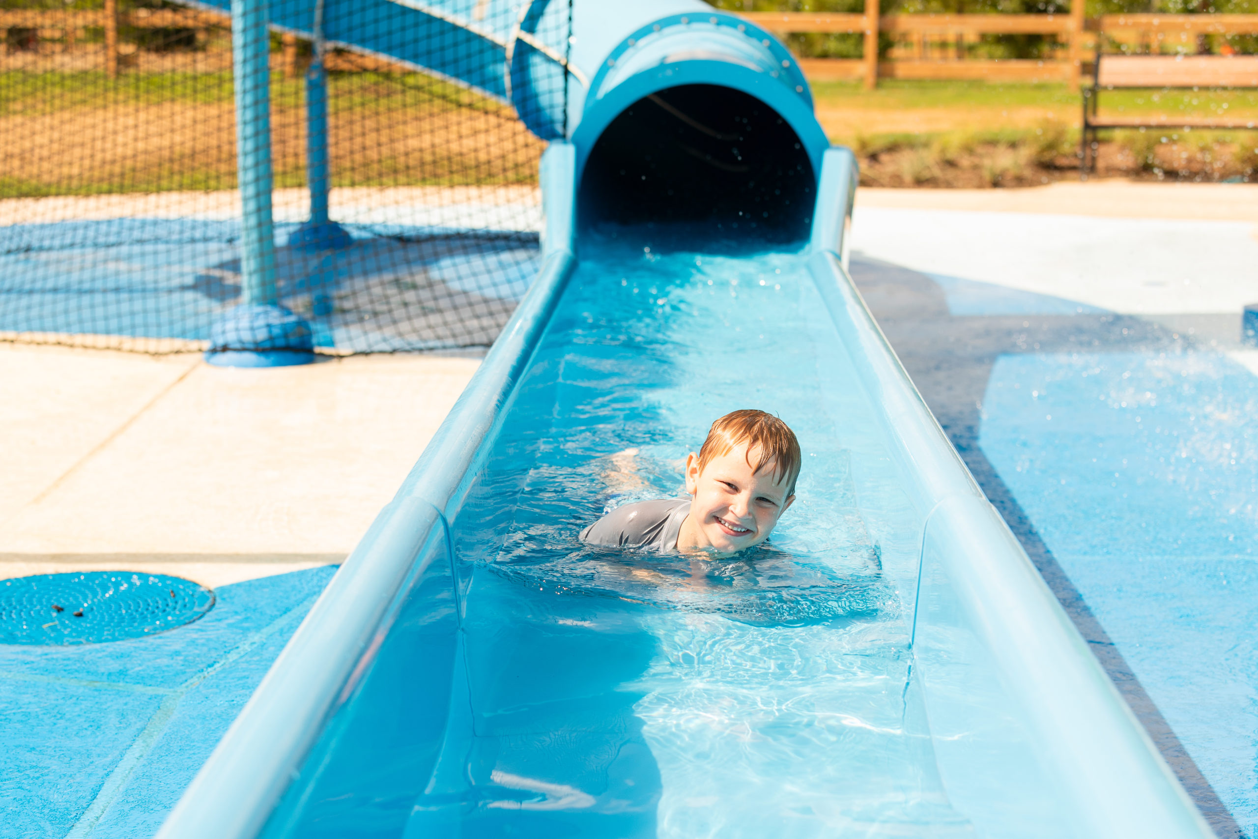 A young boy in a gray shirt enjoys the fun as he slides face down on a blue water slide at Haven Amenity Village’s outdoor water park, smiling at the camera.